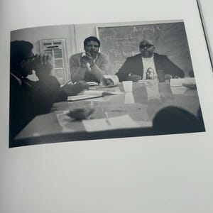 May include: Black and white photograph of three men seated at a table, engaged in a discussion. A chalkboard in the background displays the word "Agenda". The photograph is from the Black Congress, Los Angeles, 1966.