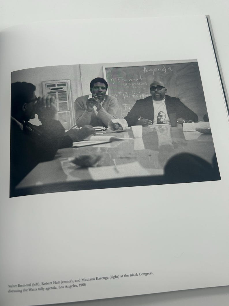 May include: Black and white photograph of three men seated at a table, engaged in a discussion. A chalkboard in the background displays the word "Agenda". The photograph is from the Black Congress, Los Angeles, 1966.