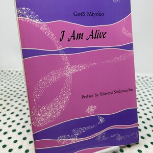 Pode incluir: Um livro intitulado "I Am Alive" de Gotō Miyoko. A capa apresenta um design com tons de roxo e rosa, com o título em preto. O prefácio é de Edward Seidensticker. O livro está sobre um suporte de metal branco.