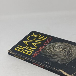 Puede incluir: Un libro de bolsillo titulado Black Brane de Michael Cisco. La portada es negra con un gr&aacute;fico de espiral amarillo, texto amarillo para el t&iacute;tulo y texto rojo para el nombre del autor. El lomo tambi&eacute;n muestra el t&iacute;tulo, el autor y la editorial Clash.