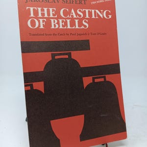 May include: A book titled "The Casting of Bells" by Jaroslav Seifert, winner of the 1984 Nobel Prize. The cover is red with black bell illustrations and text. Translated from Czech by Paul Jagasich & Tom O'Grady.