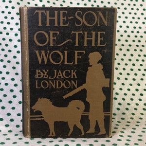 May include: The Son of the Wolf by Jack London, a vintage book cover with a black background and gold lettering. The cover features a silhouette of a person with a dog.