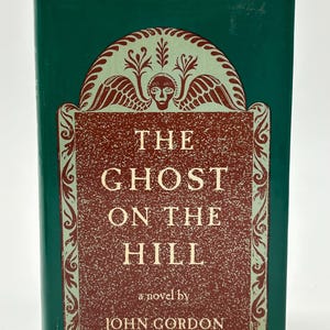 以下が含まれることがあります： ジョン・ゴードン著「The Ghost on the Hill」というタイトルのハードカバー本。表紙は濃い緑色の背景に、質感のある赤茶色の中心パネルが特徴です。装飾的なスクロール模様と、翼のある顔のイラストが表紙を飾っています。