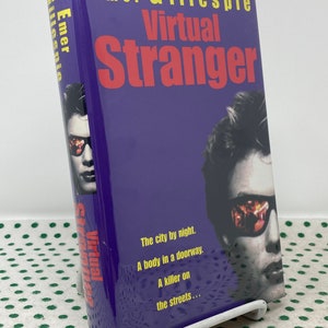 May include: A purple hardcover book titled "Virtual Stranger" by Emer Gillespie. The cover features a man wearing sunglasses. The title is in red, with the author's name in yellow. The book is on a white stand.