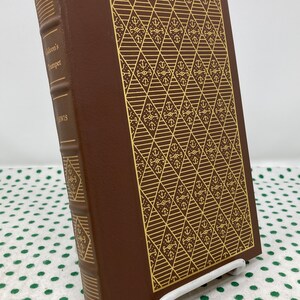 Puede incluir: Un libro encuadernado en cuero marrón con letras doradas en el lomo. El título es "Gideon's Trumpet" de Lewis. La portada presenta un patrón dorado de anclas y diamantes.