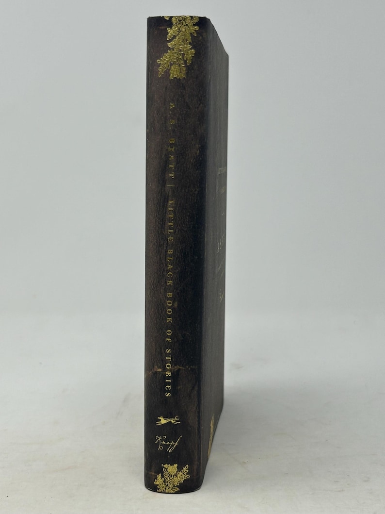 Puede incluir: Un libro marr&oacute;n oscuro titulado "Little Black Book of Stories" de A.S. Byatt. El lomo del libro presenta letras doradas y detalles florales dorados decorativos en la parte superior e inferior. El libro est&aacute; de pie sobre un fondo blanco.