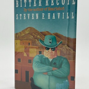 May include: A paperback book titled "Bitter Recoil" by Steven F. Havill. The cover shows a police officer in a green uniform and sunglasses, set against a desert landscape with adobe-style buildings.