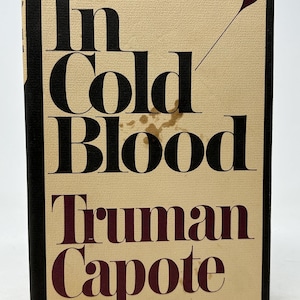May include: A hardback book titled "In Cold Blood" by Truman Capote. The cover is beige with black and burgundy lettering. A burgundy teardrop shape with a line extends from the top right corner. The book has a black spine.