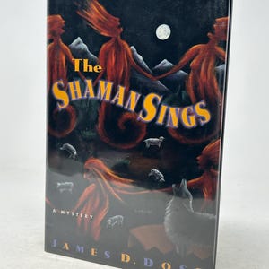 May include: A paperback book titled "The Shaman Sings" by James D. Doss. The cover features a dark background with illustrations of figures with flowing red hair, a full moon, and a wolf. The text "A Mystery" is also visible.
