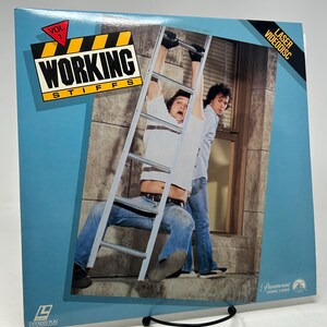 May include: A vintage Laser Videodisc for "Working Stiffs, Vol. 1." The cover features a photograph of two men, one on a ladder, with the title in bold, yellow and black text. The disc is displayed on a black metal stand against a light blue background.