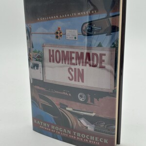 May include: A hardcover book titled "Homemade Sin" by Kathy Hogan Trocheck. The cover features a truck with the title in large red letters. The background shows a street scene with a traffic light and a yield sign.