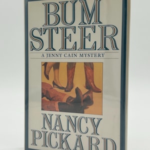 May include: A hardback book titled "Bum Steer: A Jenny Cain Mystery" by Nancy Pickard. The cover shows legs and boots, with a blue and beige colour scheme.