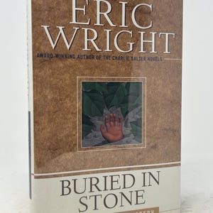 May include: A hardcover book titled "Buried in Stone" by Eric Wright. The cover features the author's name in large white letters, with the title below. A small image of a hand is in the center. The book is a Mel Pickett mystery.