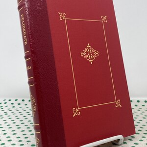 Könnte beinhalten: Ein rotes, ledergebundenes Buch mit goldener Beschriftung auf dem Rücken, die "DELIBERATE" lautet, und einem goldenen Dekor auf dem Einband.