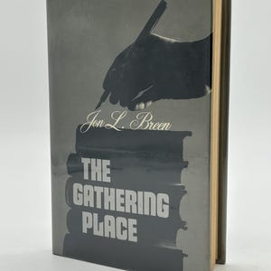 May include: A hardcover book titled "The Gathering Place" by Jon L. Breen. The cover is gray with a silhouette of a hand holding a pen above a stack of books. The book's pages have a yellowed hue.