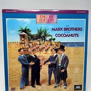 Puede incluir: Un Laserdisc vintage de "The Marx Brothers in The Cocoanuts" con una ilustración de una escena de playa. La portada presenta el título "ENCORE EDITION" y el logotipo de MCA Universal. La imagen muestra a los Hermanos Marx con trajes en una playa.