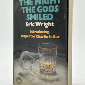 May include: A book cover with the title "THE NIGHT THE GODS SMILED" by Eric Wright. The cover features a dark background, a glass of amber liquid, and a fallen glass. The book introduces Inspector Charlie Salter.