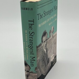 May include: A hardcover book titled "The Strangest Man" by Graham Farmelo. The book has a teal cover with the title in white and the author's name in black. The cover features a black and white image of a man.