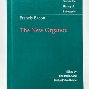 Pode incluir: Um livro de capa dura intitulado "The New Organon" de Francis Bacon. O livro tem uma capa verde-azulado com o título em rosa e o título da série "Cambridge Texts in the History of Philosophy" em branco. Editado por Lisa Jardine e Michael Silverthorne.
