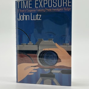 Puede incluir: Portada del libro "Time Exposure" de John Lutz. Presenta una ilustración estilizada de una cámara sobre un trípode, con manos sosteniendo la cámara. El fondo es azul oscuro con una silueta de rostro. El título y el nombre del autor son en blanco.