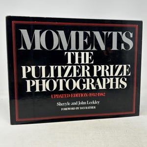 Pode incluir: Um livro de capa dura preto intitulado "MOMENTS THE PULITZER PRIZE PHOTOGRAPHS UPDATED EDITION 1942-1982" com texto branco. O livro tem uma borda vermelha e os autores são Sheryle e John Leekley.