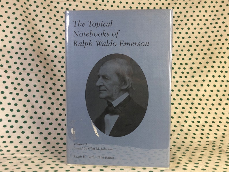 May include: The cover of a blue book titled "The Topical Notebooks of Ralph Waldo Emerson" with a black and white portrait of Ralph Waldo Emerson. The book is volume 3, edited by Glen M. Johnson and Ralph H. Orth, Chief Editor.