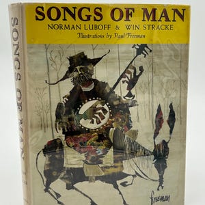Puede incluir: Un libro antiguo titulado "SONGS OF MAN" con ilustraciones de Paul Freeman. La portada presenta una imagen de estilo arte popular de una figura tocando el banjo mientras monta un burro. El lomo del libro también muestra el título.