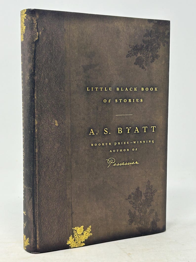 Puede incluir: Un libro de tapa dura marr&oacute;n oscuro, "Little Black Book of Stories" de A.S. Byatt. Letras doradas y dise&ntilde;os florales adornan la portada. La autora es ganadora del Premio Booker.