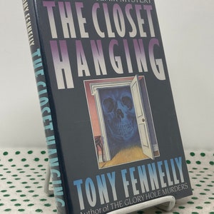 May include: A hardcover book titled "The Closet Hanging" by Tony Fennelly. The cover is dark blue with the title in large white letters. The book features an illustration of a skull in a doorway. The book is propped up on a white metal stand.