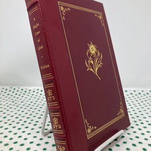 Pode incluir: Um livro encadernado em couro bordô com letras douradas e um desenho floral dourado na capa. O título do livro é "A Slight Case of Libel" de Watkins.