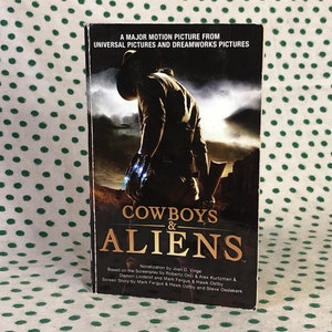 May include: A paperback book titled "Cowboys & Aliens" with a movie poster-style cover. The cover features a silhouetted cowboy figure against a desert backdrop, with the title in large, bold letters. The book is a novelization of the film.