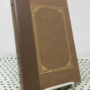 May include: A brown hardback book with gold lettering on the spine that reads "THE TRIAL OF THE U.S. 2". The book has a gold decorative border on the front cover.