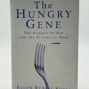 May include: A book titled "The Hungry Gene" with a light gray cover. The title is in dark blue, with the subtitle "The Science of Fat and the Future of Thin." A silver fork graphic is on the cover. The author's name, Ellen Ruppel Shell, is at the bottom.