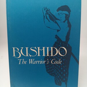 May include: A book titled "BUSHIDO The Warrior's Code" with a blue cover. The cover features a dark blue illustration of a samurai figure. The book is held up by a black stand.