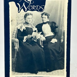 Puede incluir: Un libro antiguo titulado "In Their Own Words" con una cubierta azul oscuro. La portada presenta una fotografía en blanco y negro de dos mujeres con ropa de época. El subtítulo del libro dice "Letters from Norwegian Immigrants".