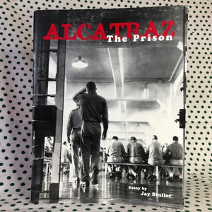 May include: Black and white photo of a book cover with the title "Alcatraz The Prison" in red letters. The cover image shows a group of men in prison uniforms walking down a hallway.