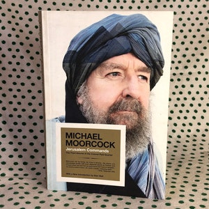 May include: A book cover featuring a portrait of a man with a beard and a blue turban. The book is titled "Jerusalem Commands" by Michael Moorcock. A gold sticker with text is on the bottom left.