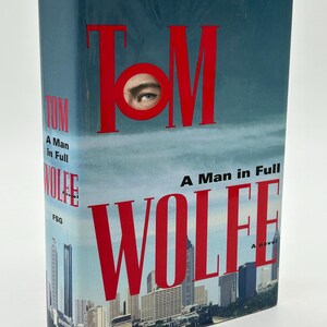 May include: A hardcover book titled "A Man in Full" by Tom Wolfe. The cover features the author's name in large red letters, with an eye incorporated into the "O". A cityscape is at the bottom.