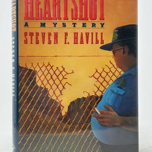 May include: A hardback book titled "HEART SHOT A MYSTERY" by Steven F. Havill. The cover shows a police officer in uniform, a chain-link fence, and a desert landscape in orange and yellow hues.
