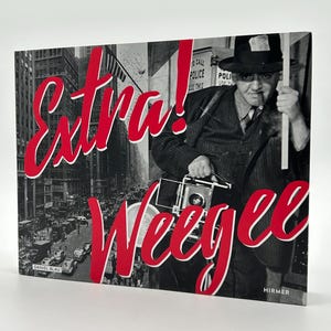 May include: A book cover featuring a black and white photograph of a man in a suit holding a camera and smoking a cigarette. The words "Extra! Weegee" are in red script across the front. The book is titled "Daniel Blau" and "Hirmer".