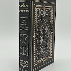 Puede incluir: Un libro de tapa dura titulado "La montaña mágica" de Thomas Mann. El libro tiene una cubierta negra con patrones y texto decorativos dorados. El lomo también presenta texto y elementos decorativos dorados. El libro está de pie.