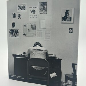 May include: Black and white photograph of a person seated at a desk, facing away from the camera. The wall behind the person is adorned with framed images and posters, including one that reads "MOVE ON OVER OR WE'LL MOVE ON OVER YOU."