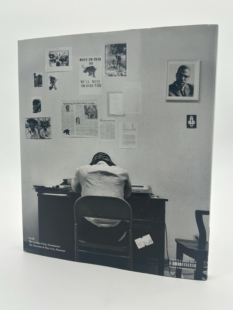 May include: Black and white photograph of a person seated at a desk, facing away from the camera. The wall behind the person is adorned with framed images and posters, including one that reads "MOVE ON OVER OR WE'LL MOVE ON OVER YOU."