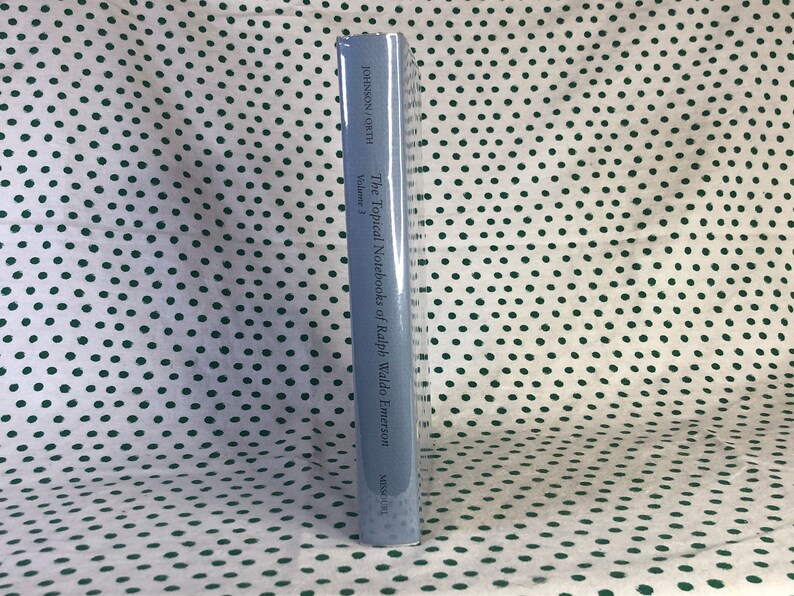 May include: The spine of a blue hardcover book titled "The Topical Notebooks of Ralph Waldo Emerson, Volume 3" by Johnson,  Couth.