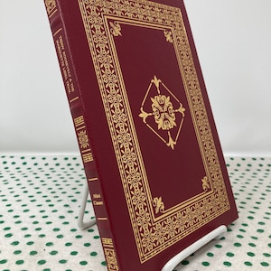 May include: A red hardback book with gold lettering on the spine. The title is "Teddy Roosevelt & The  Magnificent Bull." The author is "Mikael  Cassen".