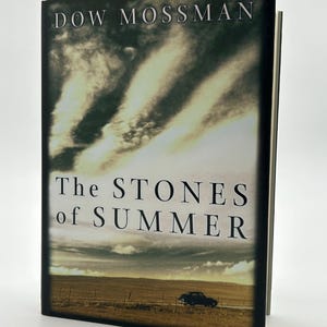 May include: A hardcover book titled "The Stones of Summer" by Dow Mossman. The cover features a landscape with a road and a car under a cloudy sky. The book is upright against a white background.