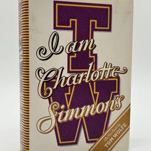 May include: A hardback book titled "I am Charlotte Simmons" by Tom Wolfe. The cover displays a large purple "T" with gold edging, and the title is in stylish script. The spine has a striped design.