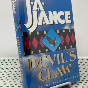 Könnte beinhalten: Ein blaues Taschenbuch mit einem weißen Adler auf dem Cover. Der Titel lautet "J.A. Jance Devil's Claw" und der Untertitel lautet "A JOANNA BRADY MYSTERY".