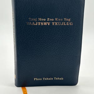 May include: A dark blue book with gold lettering on the cover. The text on the cover reads "Tzuj Moo Zoo Kws Yog VAAJTSWV TXUJLUG" and "Phoo Txhais Tshab". An orange ribbon bookmark is visible.