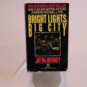 May include: A paperback book cover with a black background and a yellow title that reads "BRIGHT LIGHTS, BIG CITY" by Jay McInerney. The cover features a photo of a city street at night with a man walking in the foreground.